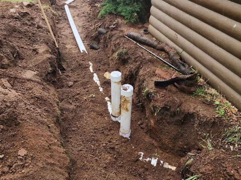 Excavated trench beside a log-sided house with new white PVC sewer pipe and cleanouts, excavator in the background
