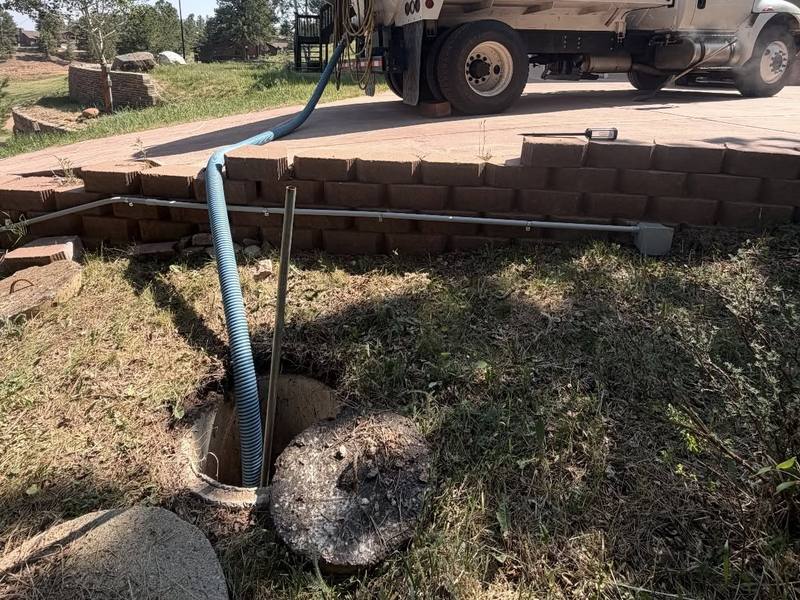 TCS pump truck parked on a driveway by a house, with its hose running down to an uncovered septic tank
