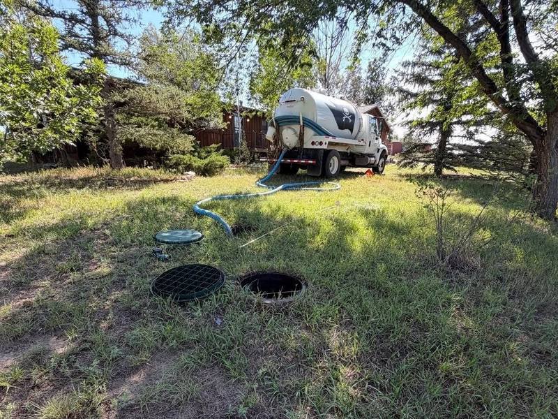 TCS pump truck parked behind two open septic tank lids in a grassy yard, hose laid out to the tank